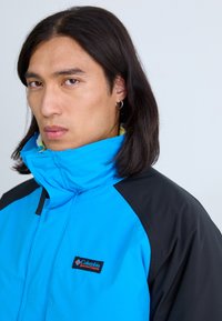 Blau- und schwarze Außenjacke mit hohem Kragen, Reißverschluss und sichtbarem Branding. Das Material wirkt glatt mit einer kontrastierenden Textur.