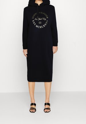 Granatowa sukienka dresowa z kapturem i nadrukiem na przodzie, z długimi rękawami i długością do połowy ud. Noszona z czarnymi sandałami na paski.