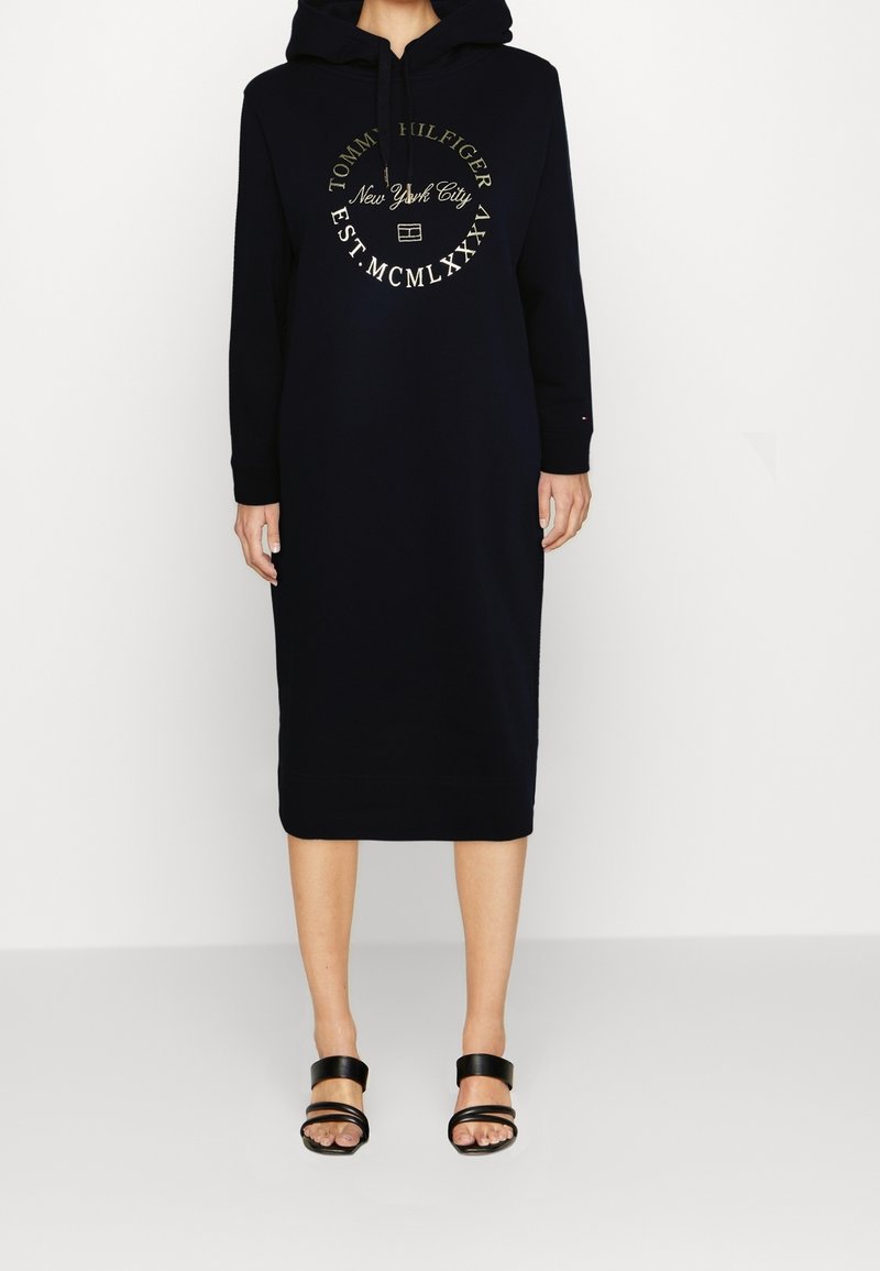 Robe sweat à capuche navy avec un imprime de logo sur le devant, manches longues et coupe mi-longue. Portée avec des sandales noires à lanières.