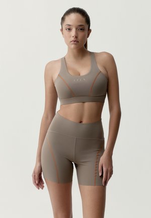 Žena v béžovom športovom krátkom topu a krátkych cyklistických šortkách s vysokým pásom a oranžovými švovými detailmi, stojaca pred jednoduchým svetlým pozadím.