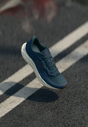 Încălțăminte sportivă albastră și albă în aer, deasupra asfaltului texturat cu linii albe vopsite și resturi mici împrăștiate dedesubt.