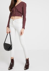 Crop top bordeaux à pois avec détails de boutons, associé à un jean skinny blanc et un sac à main noir. Bottines marron brillantes avec des accents métalliques.