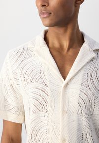 Witte kanten blouse met een golvenpatroon, korte mouwen, een kraag en knoopsluiting. De gedetailleerde textuur voegt diepte toe aan de lichte stof.