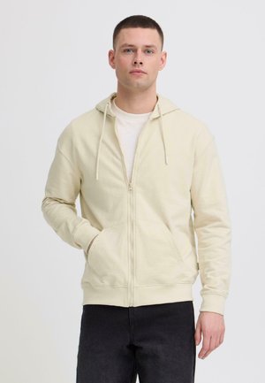 Ung mand iført en cremefarvet hættetrøje med lynlås og sorte bukser, stående med den ene hånd i lommen mod en ensfarvet hvid baggrund.