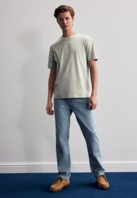Hellgrünes Baumwoll-T-Shirt mit kurzen Ärmeln, kombiniert mit hellblauen Jeans und beigen Clogs, stehend vor einem einfarbigen Hintergrund.