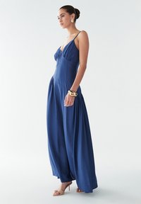 Zila maksi kleita ar plānām lencēm, pieguļošu augšdaļu un plūstošu svārku. Gluda auduma, potīšu garuma, papildināta ar bēša krāsas papēžu sandales un zelta rokassprādzi.