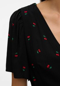 Schwarzes Kleid mit kurzen Puffärmeln, V-Ausschnitt und einem Kirschmuster in Rot und Grün. Weicher Stoff mit Raffung details an der Schulter.