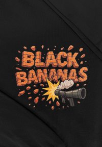 Zwart T-shirt met opvallende oranje tekst "BLACK BANANAS" met explosieve afbeeldingen, een kanon en losbrekende stenen, tegen een textuurrijke achtergrond.