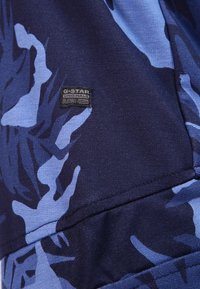 Tessuto blu a motivo camouflage con sfondo blu scuro; include una patch nera con il logo G-Star. Texture liscia, con cuciture dettagliate visibili.
