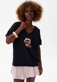 Zwarte T-shirt met korte mouwen en een V-hals, versierd met een cartoonkatgrafisch ontwerp. Gecombineerd met een lichtroze gelaagde rok. Gladde stofstructuur.