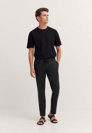Mann steht gekleidet in ein schwarzes kurzärmliges T-Shirt, schwarze Jogginghosen mit Kordelzug und braune Sandalen vor einem schlichten hellen Hintergrund.