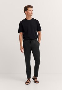 Schwarzes Kurzarmhemd, lockere Passform. Schwarze Hose mit Kordelzugbund und seitlichen Taschen. Braune Sandalen mit zwei Riemen und flacher Sohle.