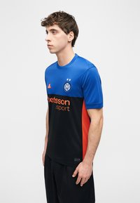 Νέος άνδρας με μπλε και μαύρη αθλητική φανέλα με πορτοκαλί πλευρικές π ινακίδες και λογότυπα, στέκεται προφίλ σε λευκό φόντο.