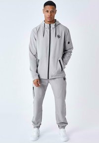 Chaqueta atlética gris con capucha y logotipo negro, combinada con pantalones de chándal grises a juego que tienen bolsillos con cremallera y puños elásticos, llevada con zapatillas blancas.