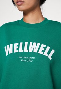 Grön sweatshirt med vit text som läser "WELLWELL" ovanför "inte intresserad av sport sedan 2002." Gjord av mjukt tyg med rund halsringning.