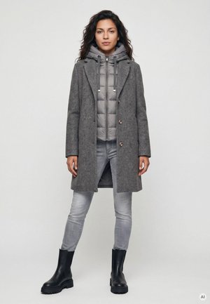 Frau mit dunkel lockigem Haar, die einen grauen Wollmantel über einer grauen Steppjacke, hellgraue Jeans und schwarze Stiefel trägt und vor einem einfarbigen Hintergrund steht.