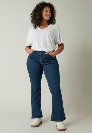 Witte V-hals T-shirt gecombineerd met hooglopende donkerblauwe flare jeans. Model draagt witte sneakers, casual fit met zichtbare zakken.