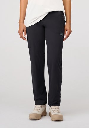 Osoba nosząca czarne spodnie o prostych nogawkach z boczną kieszenią na zamek i beżowe buty trekkingowe, stojąca na jednolitym jasnym tle.