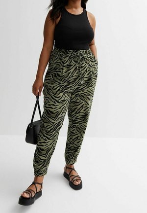 Femme portant un haut noir sans manches, un pantalon imprimé zèbre vert et noir, des sandales à plateforme noires et tenant un petit sac à main noir.