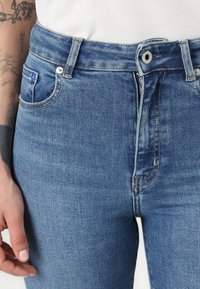 Mėlyni džinsai su aukštu liemeniu, klasikinio penkių kišenių dizainu ir užtrauktuko priekyje su mygtuku. Minkšta tekstūra ir šiek tiek išblukusi apdaila.