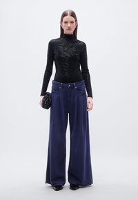 Μαύρο τοπ με παγιέτες και ψηλό λαιμό, σε συνδυασμό με υπερμεγέθη μπλε σκούρα παντελόνια με φαρδιά μπατζάκια. Το μοντέλο κρατάει μαύρο φάκελο-τσάντα· κομψή υφή και γυαλιστερό φινίρισμα.