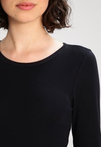 Schwarzes Langarm-Oberteil mit runder Halslinie, aus einem glatten, elastischen Stoff gefertigt. Nahaufnahme, die sich auf den oberen Brust- und Halsbereich konzentriert.