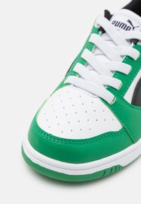 Sneaker sportivo bianco e verde con punta perforata, parte superiore in pelle testurizzata, dettagli neri e lacci bianchi, dotato di una spessa suola in gomma.