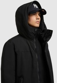 Hombre vestido con chaqueta con capucha negra y gorra de béisbol negra de los New York Yankees, mirando hacia la derecha contra un fondo gris claro.