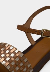 Hnědá proplétaná kožená sandála s kovovým akcentem, nastavitelným páskem kolem kotníku a plochou podrážkou. Má texturovaný povrch a elegantní design.