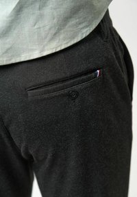 Pantalon noir sur mesure avec une poche arrière boutonnée affichant un petit onglet en tissu tricolore, porté avec une chemise vert clair rentrée.