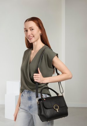 Sac à main en cuir noir au design structuré, avec ferrures rondes et une bandoulière amovible. Le mannequin porte un haut vert olive et un jean en denim clair.