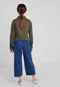 Oliwkowa krótka kurtka z epoletem, w połączeniu z niebieskimi dżinsami o szerokich nogawkach z surowym brzegiem i dwiema tylnymi kieszeniami.