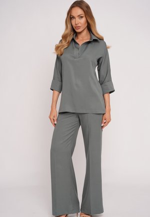 Kobieta ubrana w luźny szary top z kołnierzykiem i rękawami 3/4 oraz dopasowane szerokie spodnie, stojąca na jednolitym tle.