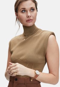 Brauner, gerippter ärmelloser Oberteil mit Stehkragen; verfügt über geschichtete Texturen und ein enganliegendes Design. Die Accessoires umfassen ein goldenes Armband und eine Uhr.