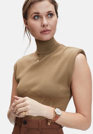 Bruine geribbelde mouwloze top met een hoge hals; bevat gelaagde texturen en een aansluitend ontwerp. Accessoires zijn een gouden armband en een horloge.