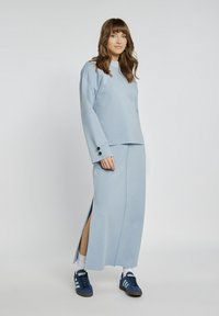 Jasnoniebieska bluza z długim rękawem i akcentami z guzików, zestawiona z dopasowaną długą spódnicą z rozporkami, noszona z niebieskimi trampkami.