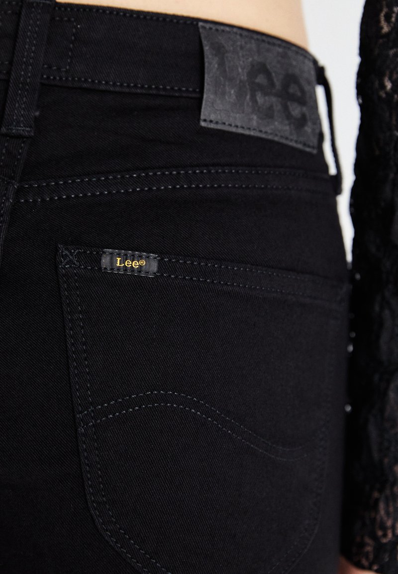 Jean en denim noir avec un design de couture visible, un patch en cuir sur la taille avec un logo en relief, et une petite étiquette de marque sur la poche.