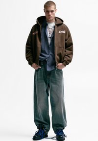 Brun hættetrøje med hvid tekst, lagdelt over en blå ternet skjorte, båret med løse grå jeans og sorte støvler med blå snørebånd.