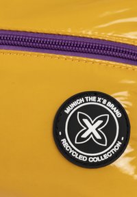 Bolsa amarilla brillante con cremallera morada y un parche circular negro con texto blanco que dice "COLECCIÓN RECICLADA". Líneas limpias y textura suave.