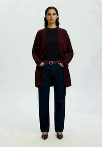 Cardigã malhado em burdeos, de frente aberta, usado sobre uma blusa preta. Combinado com jeans de perna reta azul escuro e sapatos de salto agulha burdeos.