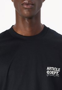 Juodas medvilninis marškinėlis su apvaliu iškirpimu ir trumpomis rankovėmis, kairėje krūtinės pusėje spausdinta balta "ARTICLE DEPT." užrašas.