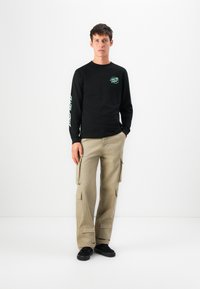 Zwarte langetermijn shirt met groene grafische print, gecombineerd met beige cargobroeken met zijzakken en zwarte sneakers. Eenvoudig, functioneel ontwerp.