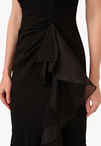 Abito nero con strati di volant sui lati. Realizzato in tessuto liscio, presenta una silhouette aderente e un accentuato volante trasparente.