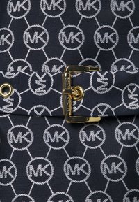 Námořnická tkanina s opakujícím se hexagonálním vzorem bílých "MK" log, zajištěná zlatou kovovou sponou a ozdobnými kroužky.
