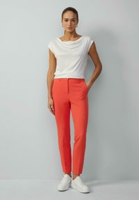 Weißes Drapiertop kombiniert mit schmalen korallenfarbenen Hosen und weißen Sneakers. Das Outfit zeichnet sich durch ein minimalistisches Design mit glatten Texturen aus.