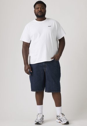 Mand stående iført hvid Levi's t-shirt, mørkeblå denimshorts, hvide sokker og hvide sneakers mod en ensfarvet lys baggrund.