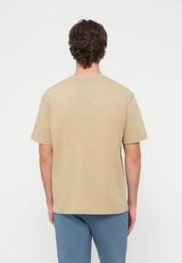 Béžové bavlnené tričko s klasickým okrúhlym výstrihom a krátkymi rukávmi, zobrazené zozadu, v kombinácii so svetlomodrými nohavicami.