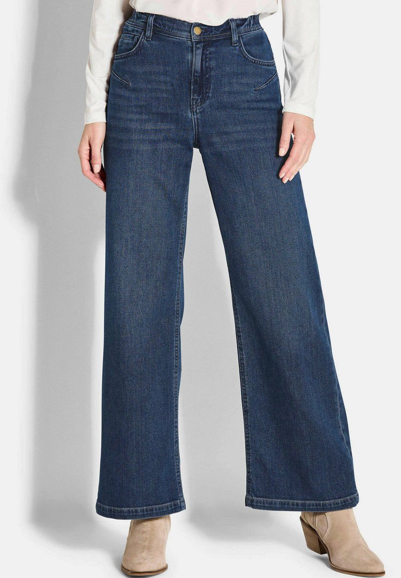 Jeans en denim à jambes larges de couleur bleu foncé avec une taille haute, présentant un design à cinq poches, des coutures discrètes et un ourlet propre. Complétés par des bottines beiges.