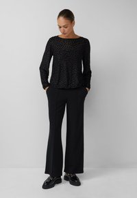 Vrouw in een zwarte gestructureerde lange mouwen top, zwarte wijde broek en zwarte glanzende loafers, staand tegen een effen lichte achtergrond.