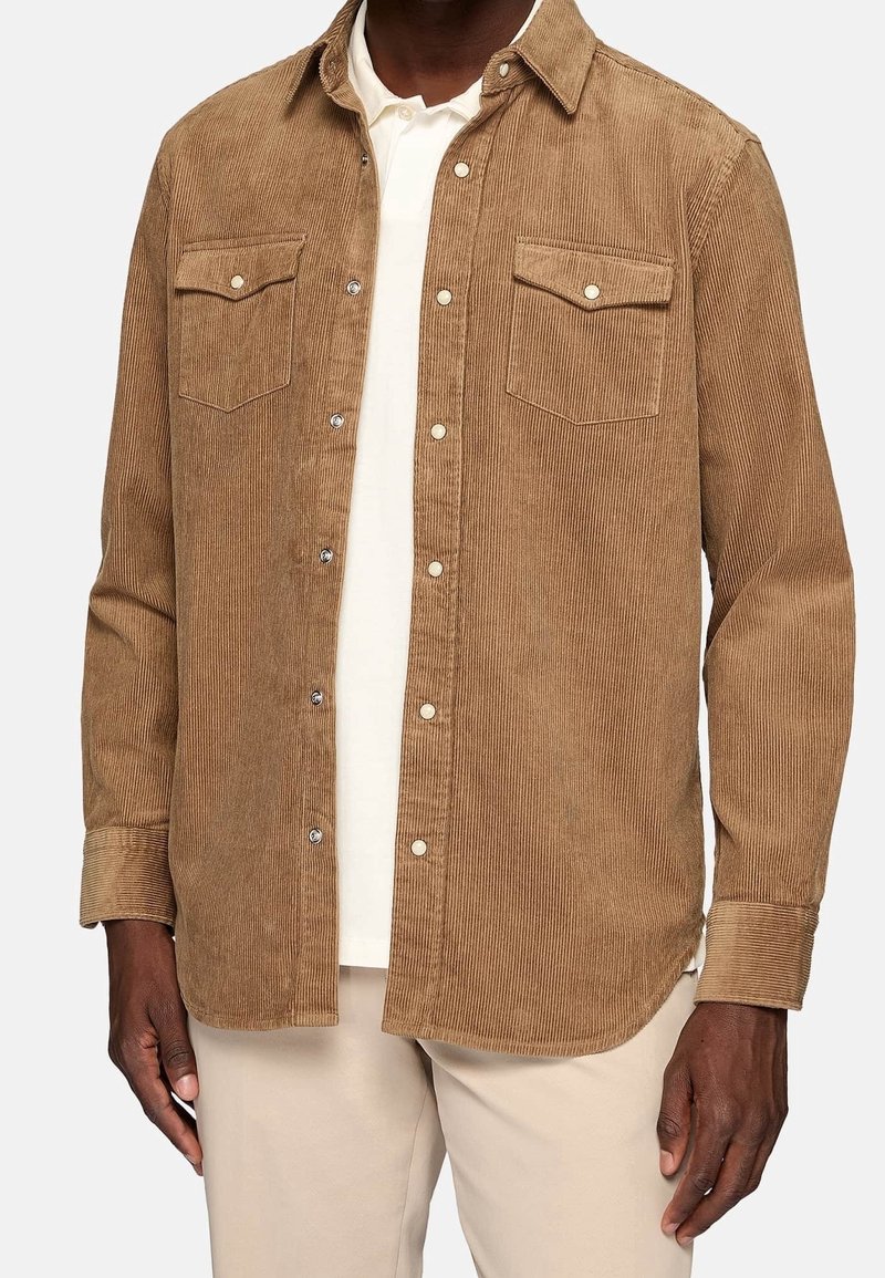 Bruine corduroy shirt met lange mouwen, knoopsluiting aan de voorkant, twee borstzakken en zichtbare ribbelstructuur. Draag het over een witte laag.